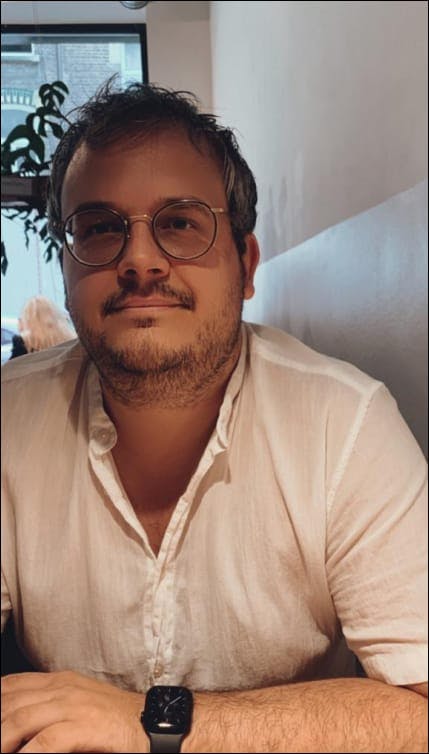 Me wearing glasses, a white blouse and having a very short beard, sitting in a coffee bar.